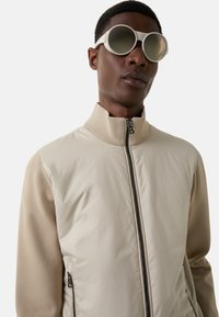 Man wearing oversized round beige sunglasses and a beige zip-up jacket with textured sleeves and smooth front panel.