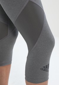 Detailní záběr na šedé sportovní legíny s síťovými panely na stehnech a logem Adidas blízko kolena, nošené na ohnuté noze na jednolitém pozadí.