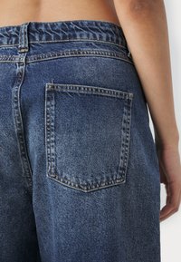 Blå denim jeans med en afslappet pasform, der har en elastisk talje, baglomme og synlig søm. Teksturen er glat med en falmet finish.