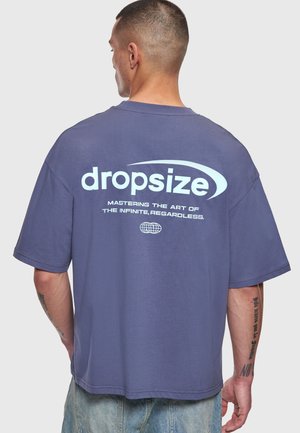 Bărbat cu păr scurt purtând un tricou lejer mov cu textul „dropsize” și un slogan pe spate, asortat cu pantaloni scurți din denim albastru deschis.