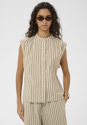 Žena s kučeravými vlasmi nosiaca hnedé slnečné okuliare, zlaté kruhové náušnice, bezrukávové béžové a olivovo pruhované zapínacie tričko a zladené nohavice.