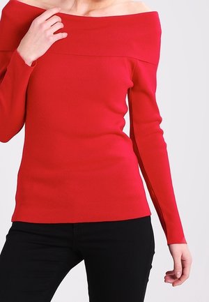 Maglione rosso off-shoulder con maniche lunghe, texture a coste e design aderente. Abbinato a pantaloni neri, con una mano visibile sulla manica.