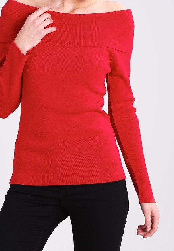 Maglione rosso off-shoulder con maniche lunghe, texture a coste e design aderente. Abbinato a pantaloni neri, con una mano visibile sulla manica.