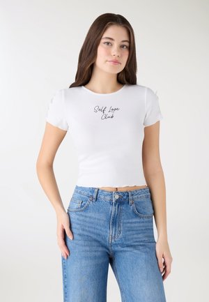 Giovane donna con lunghi capelli castani che indossa un top corto bianco con la scritta "Self Love Club" e jeans a vita alta blu, in piedi contro uno sfondo chiaro.