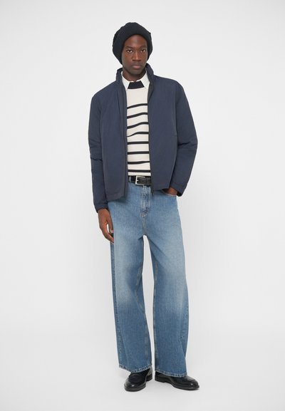 Ναυτικό πλούσιο μπουφάν πάνω από μια ριγέ ζακέτα, συνδυασμένο με φαρδιά ανοιχτό μπλε τζιν και μαύρες μπότες. Ένα μαύρο πλεκτό καπέλο ολοκληρώνει το σύνολο.