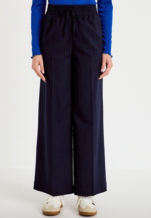 Personne portant un pantalon large à rayures bleu marine foncé avec taille élastique et cordon noir, associé à des baskets beige et blanches.