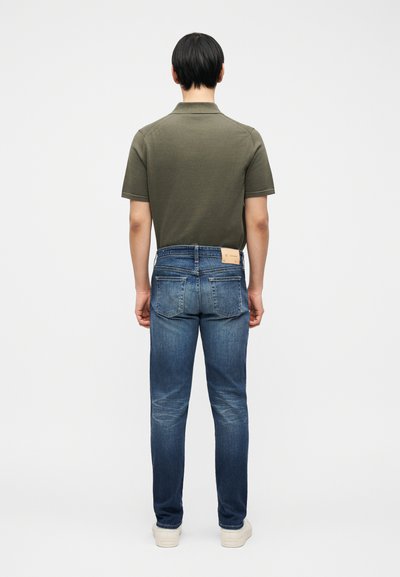 Άτομο με κοντά μαύρα μαλλιά που στέκεται γυρισμένο πλάτη, φοράει μια πουκάμισο πόλο ελιάς, μπλε τζιν και λευκά αθλητικά πάνω σε απλό φόντο.