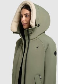 Olivovo zelená bunda s kapucňou, podšitá krémovým fleecom, so čiernymi kovovými doplnkami, predným zipsom a bočnými vreckami. Výrazné logo na hrudi.