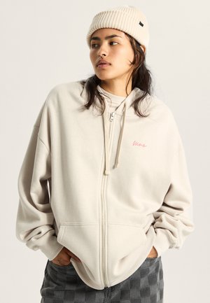 Light beige zip-up hoodie with a relaxed fit, featuring a small pink logo and kangaroo pockets, paired with a ribbed beige beanie.