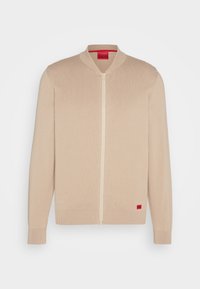 Pull beige à fermeture éclair en maille avec poignets, col et ourlet côtelés, comportant un petit patch rectangulaire rouge près de l'ourlet inférieur.