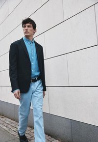 Sort blazer over en lys blå skjorte med knapper, parret med lys blå jeans og sorte sko, der går mod en struktureret lys væg.
