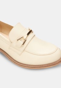 Mocassino in pelle color crema con un accentuazione in metallo dorato, caratterizzato da una punta arrotondata e una suola in materiale marrone testurizzato.