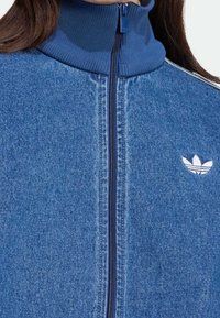 Chaqueta de mezclilla con cremallera y cuello acanalado azul, que presenta un logotipo blanco en el pecho y una cremallera azul oscura. Tejido texturizado con diseño sin costuras.
