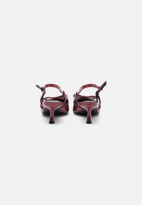Ζευγάρι γυαλιστερών μπορντό τακουνιών slingback με μεσαίο ύψος και λεπτά τακούνια, προσανατολισμένα από το πίσω μέρος σε λευκό φόντο.