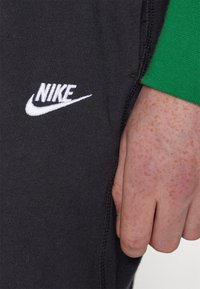 Calças de moletom pretas em algodão com o logótipo "Nike" bordado em branco. Primeiro plano mostra o tecido texturizado e a costura visível. A manga de uma camisola verde está parcialmente visível.