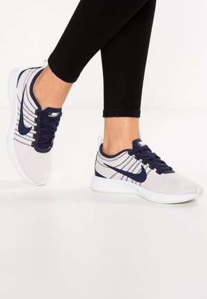 Osoba nosząca białe i granatowe buty do biegania Nike oraz czarne spodnie na jednolitym białym tle.