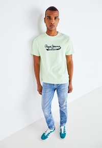 Gaiši zaļa kokvilnas T-kreklu ar melnu "Pepe Jeans" logo, saskaņota ar gaiši ziliem nolietotiem džinsiem un teal krāsas sporta apaviem. Vienkāršs dizains, brīvs piegriezums.