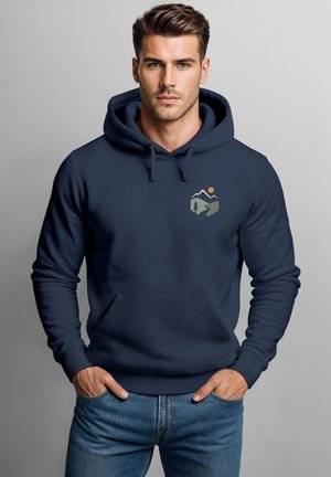 Mann mit heller Haut und gestyltem braunem Haar, der einen marineblauen Hoodie mit kleinem Berg- und Sonnengrafik trägt, die Hände in den Taschen, vor grauem Hintergrund.