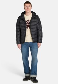 Man i en svart pufferjacka över en beige skjorta med en trädgrafik, blå jeans och svarta stövlar, stående mot en vit bakgrund.