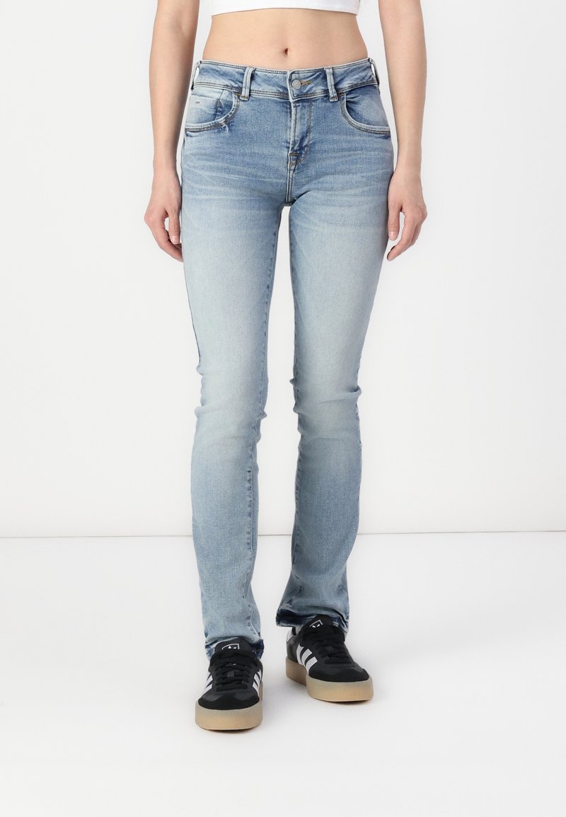 Femme portant un jean skinny bleu clair et des baskets noires avec des bandes blanches, debout devant un fond blanc.