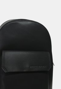 Mochila de cuero negro con parte superior redondeada, bolsillo frontal con tapa y logo en relieve "ARMANI EXCHANGE". Textura suave con un patrón sutil.