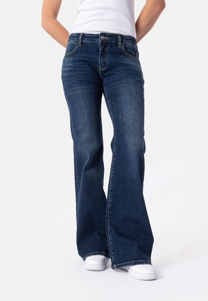 Person iført mørkeblå vidde jeans og hvide sneakers, stående mod en ensfarvet lys baggrund.