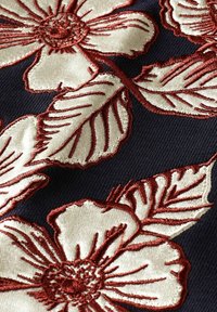 Wyhaftowany wzór florystyczny z kremowymi kwiatami i ciemnoczerwonymi konturami na ciemnoniebieskiej teksturowanej tkaninie, ukazujący szczegółowe płatki i liście.