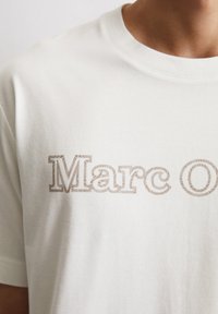 Weißes Baumwoll-T-Shirt mit dem Text "Marc O" in einem braunen Texturmuster. Kurze Ärmel und runder Ausschnitt.