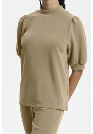 Femme portant un ensemble beige composé d’un haut et d’un pantalon assortis, haut à col haut et manches bouffantes aux coudes.