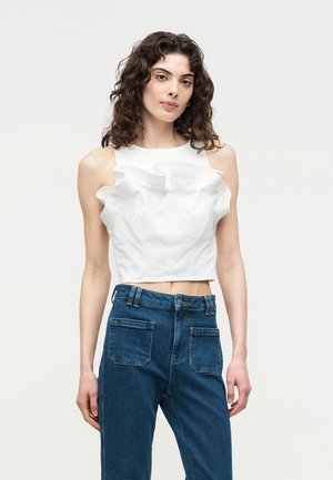 Kobieta z kręconymi, ciemnymi włosami, ubrana w białą, bezrękawową, marszczoną crop top i wysokie, niebieskie dżinsy, stojąca na jednolitym tle.