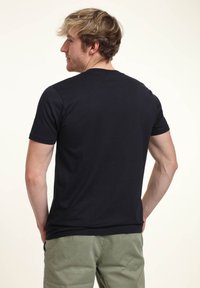 Joven con cabello corto y ondulado, llevando una camiseta negra lisa y pantalones verde claro, de pie de espaldas a la cámara.