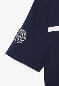 Polo de color azul marino con textura acanalada, presenta una franja blanca y un parche redondo del Club de Tenis Lacoste en la manga izquierda.