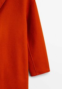Cappotto in misto lana arancione con una consistenza morbida, caratterizzato da una vestibilità ampia e una manica larga e inclinata. Senza chiusure o dettagli aggiuntivi visibili.