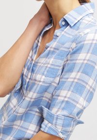 Modrá a bílá kostkovaná košile na knoflíky s límečkem, s vyhrnutými rukávy a náprsní kapsou. Materiál působí lehce a má strukturovaný vzhled.