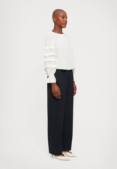 Femme avec la tête rasée portant un chemisier blanc à manches volantées, un pantalon large à rayures noires, et des escarpins blancs se tenant sur le côté.