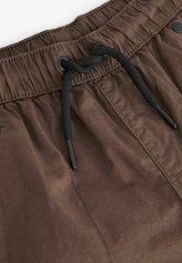 Braune Badeshorts mit elastischem Bund, ausgestattet mit schwarzem Kordelzug und metallischen Akzenten. Der Stoff wirkt glatt und hat einen subtilen Glanz.