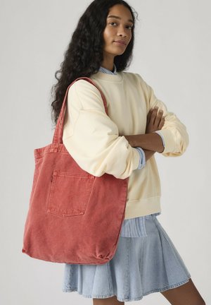 Donna con capelli lunghi e ricci che indossa una felpa color crema e una gonna di jeans, con una grande borsa tote in velluto a coste rossa sulla spalla.