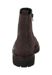 Botín de tobillo de ante marrón oscuro con tirador y suela gruesa negra, visto desde la parte trasera sobre un fondo blanco.