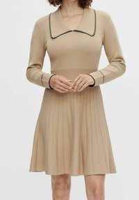 Neule beige mekko pitkähihaisilla, leveällä kauluksella ja mustilla yksityiskohdilla. Mekkossa on laskostettu hame, joka luo levenevän siluetin.