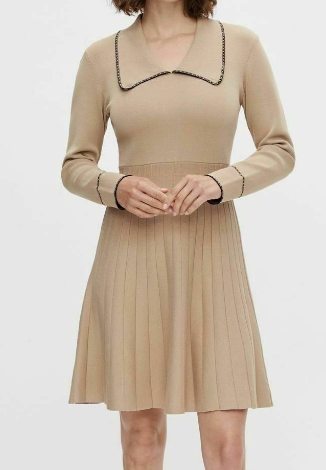 Neule beige mekko pitkähihaisilla, leveällä kauluksella ja mustilla yksityiskohdilla. Mekkossa on laskostettu hame, joka luo levenevän siluetin.