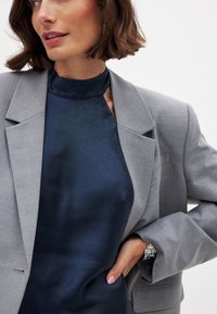 Grau, taillierter Blazer über einem dunkelblauen, seidenartigen Oberteil mit hohem Kragen. Silberne Uhr, die am Blazerärmel sichtbar ist. Subtile Strukturen in den Materialien.