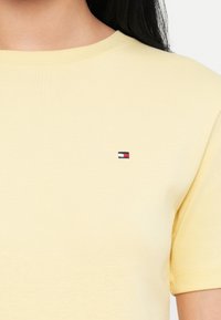 Gul t-skjorte med korte ermer laget av bomull, rund hals og en liten brodert logo i rød, hvit og blå på brystet.