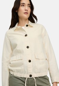 Woman wearing a cream buttoned jacket with two front flap pockets and olive green pants, looking slightly upward.