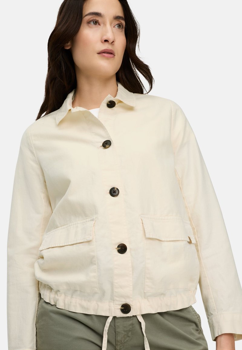 Woman wearing a cream buttoned jacket with two front flap pockets and olive green pants, looking slightly upward.