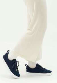 Marineblaue Canvas-Sneaker mit weißen Gummisohlen, Schnürdesign und Schlaufenakzenten, getragen mit weißen gerippten Socken und weiten cremefarbenen Hosen.