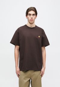 Brown cotton t-shirt with short sleeves and a crew neck, featuring a small orange logo on the chest. Regular fit, simple design.
