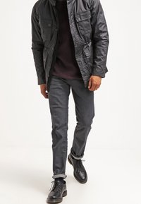 Czarna, błyszcząca, teksturowana kurtka z wieloma kieszeniami, noszona na bordowym swetrze. Ciemnoszare dopasowane jeansy i czarne buty sznurowane dopełniają stylizację.