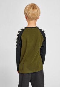 Barn med kort blondt hår iført en olivengrøn og sort langærmet skjorte med hvide chevron-striber, stående med ryggen mod en ensfarvet baggrund.