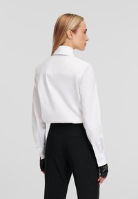 White cotton shirt with a structured collar, fitted back, and long sleeves, paired with black high-waisted trousers and fingerless gloves.