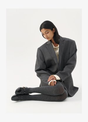 Blazer grigio oversize sopra una camicia chiara, abbinato a collant grigi con texture e scarpe nere lucide con un dettaglio a fibbia.